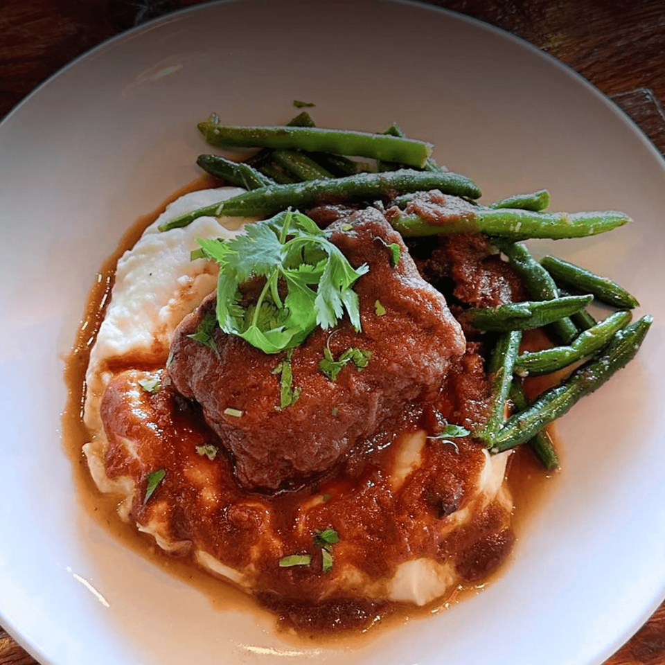 Braised short rib with mashed potatoes, gravy, and green beans on a white plate.