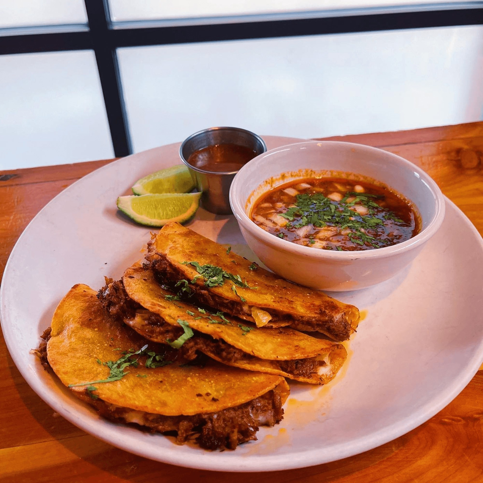 Birria tacos with a side of consommé, lime wedges, and a small cup of sauce on a white plate.