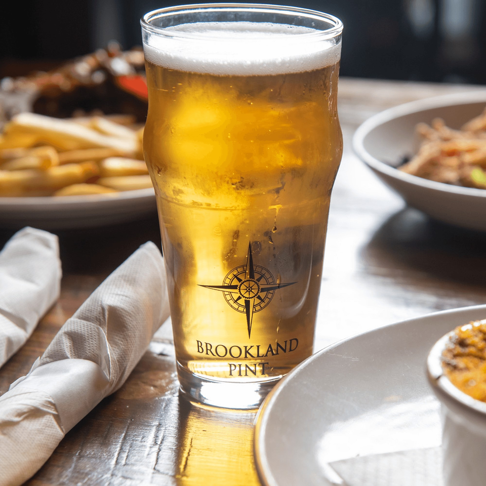 A Brookland Pint glass of golden beer with foam, alongside fries and other dishes on a wooden table.