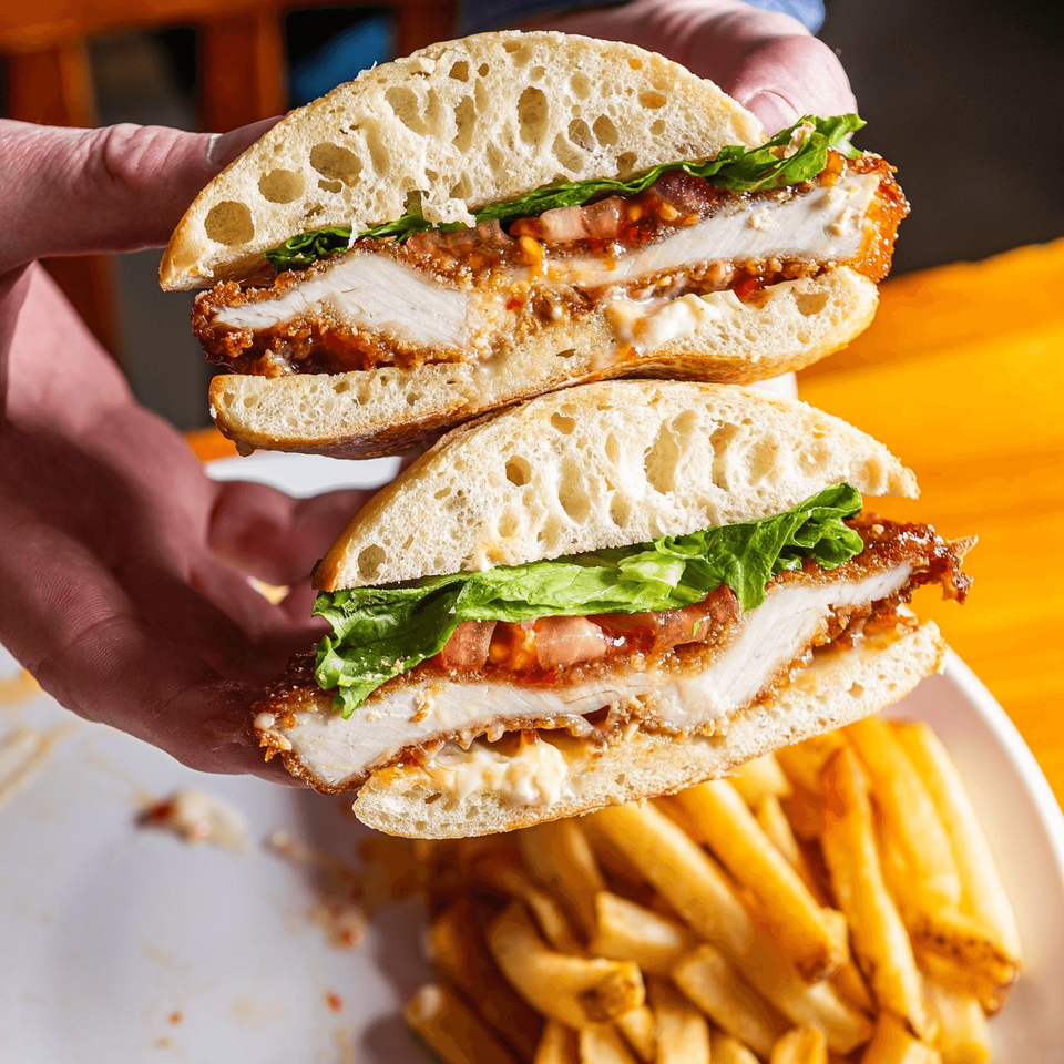 Hands holding a Brookland Pint fried chicken sandwich, cut in half, with lettuce, tomato, and fries.